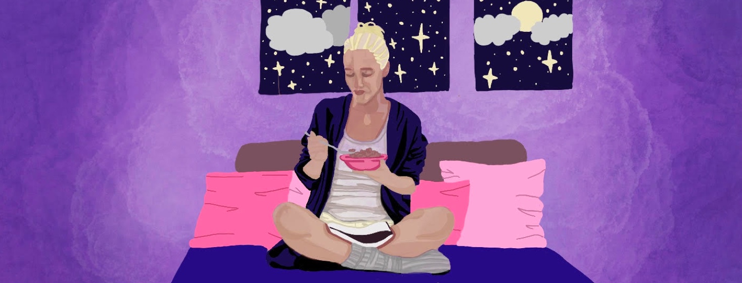 A woman at night sits on her bed eating cereal and reading a book in her lap